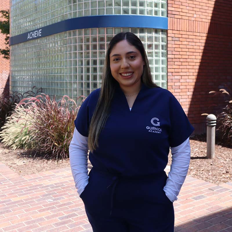 young woman in scrubs in front of hospital, smiling